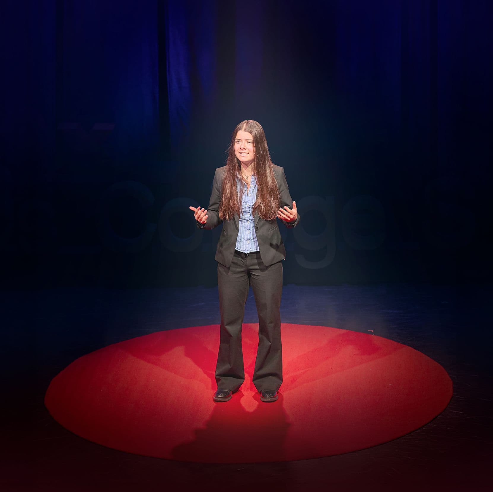 Woman in suit on stage, illuminated by red spotlight, gesturing during a presentation.