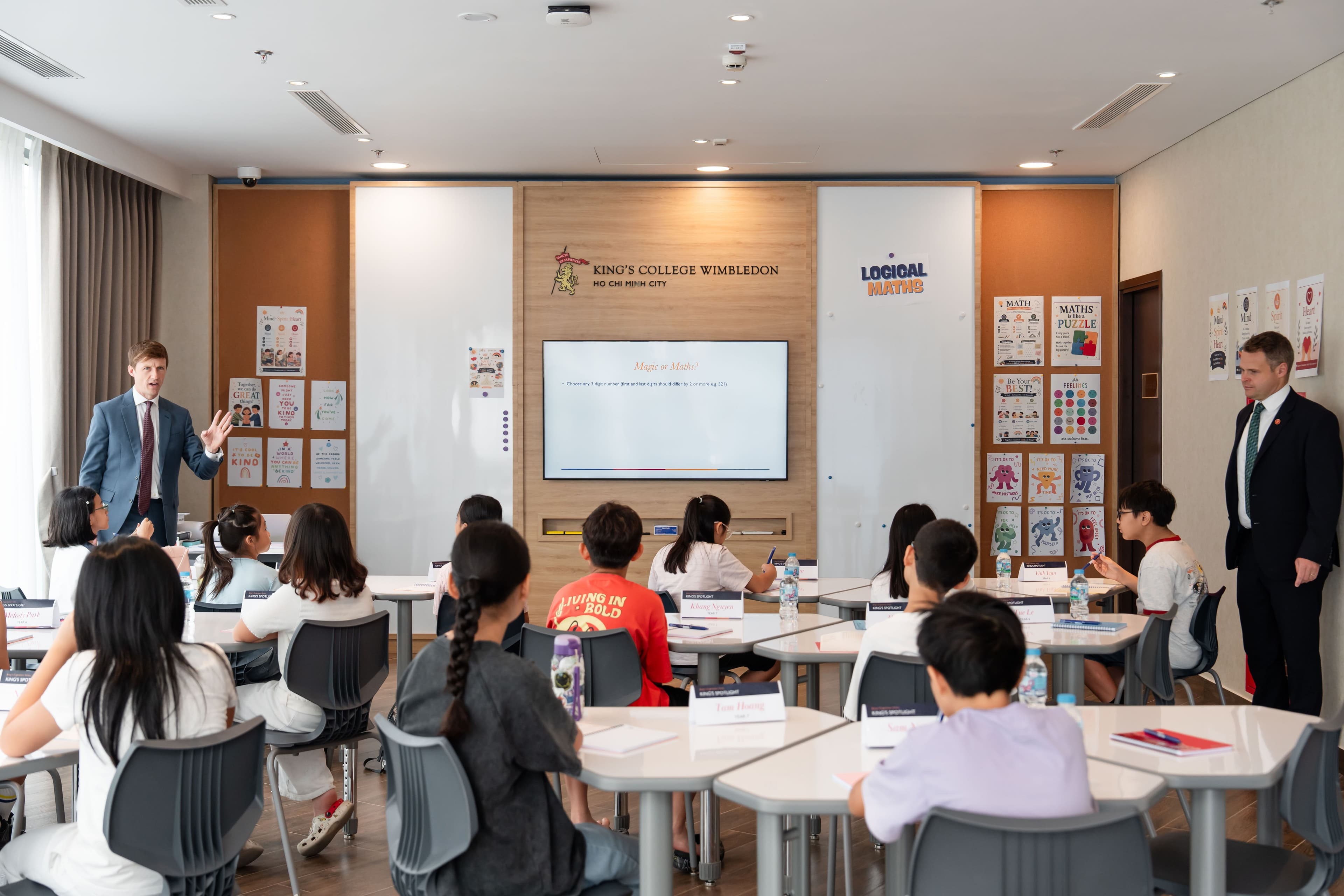 Two male instructors teach students in a modern classroom with a large screen.