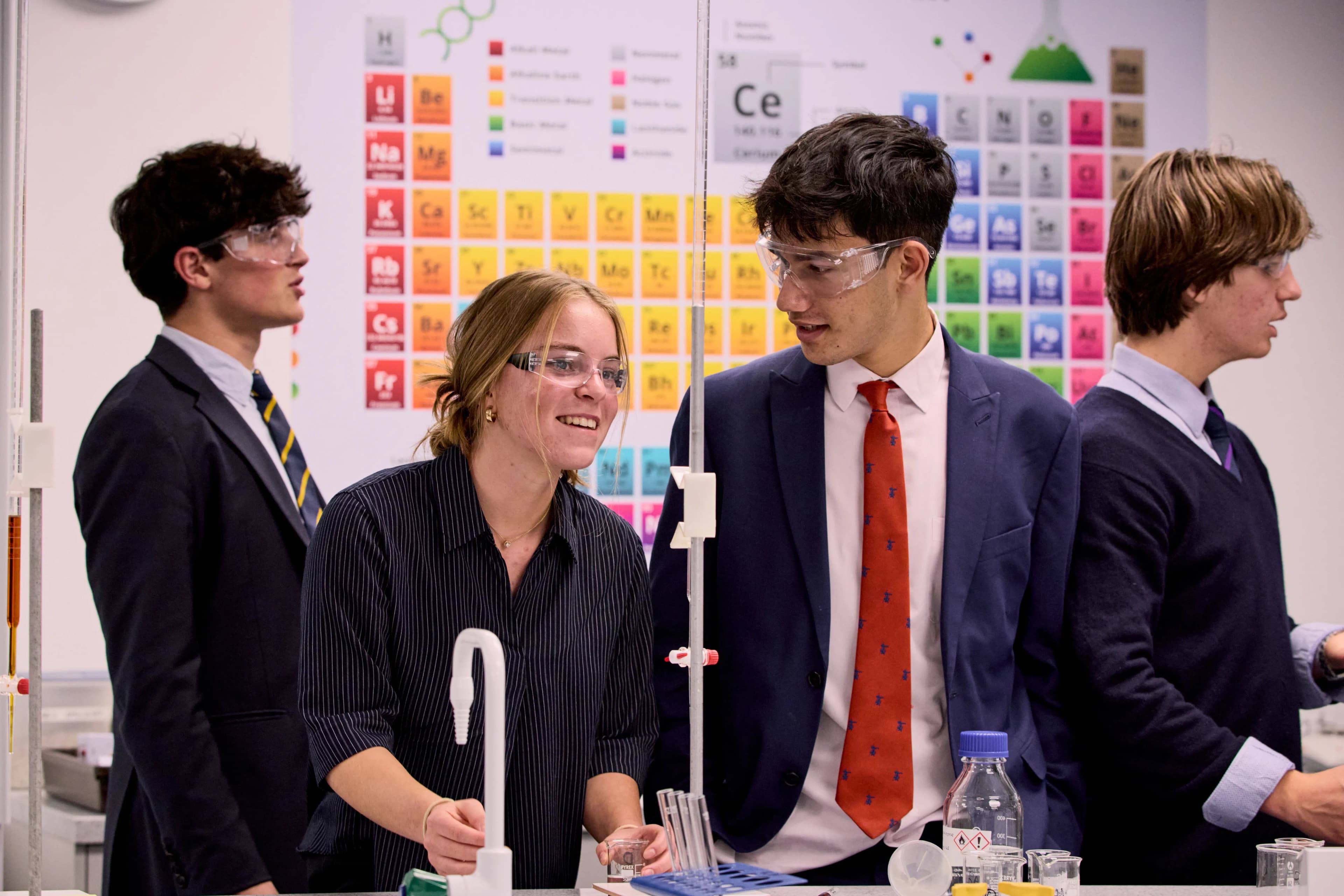 Four students wearing safety glasses conduct an experiment in a science lab with a periodic table.
