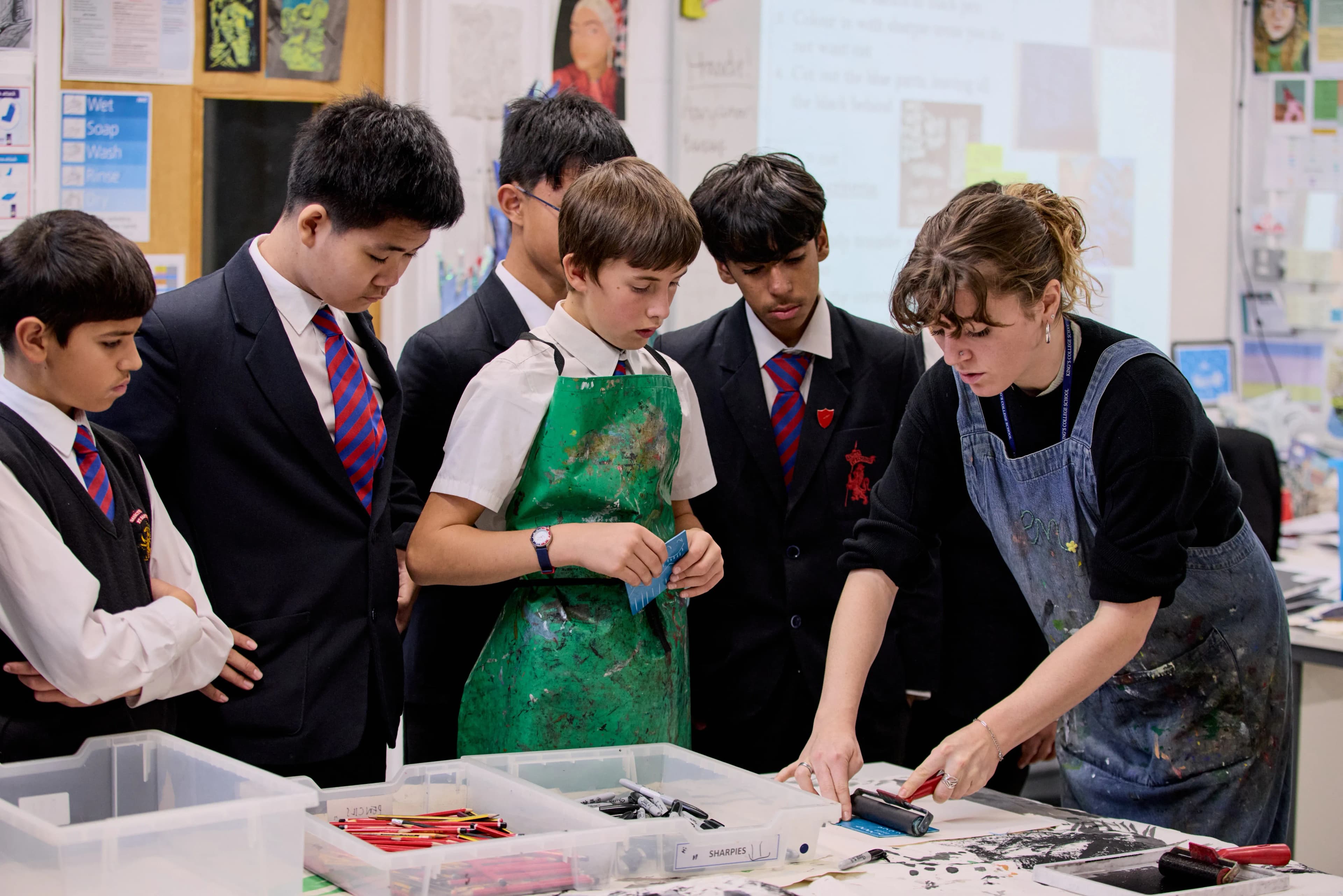 Teacher demonstrates printmaking technique to five male students in an art classroom.