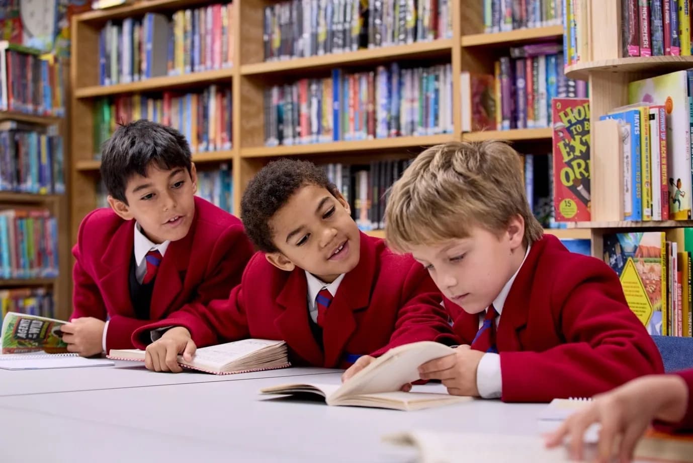 Three young students in red uniforms read books together at a library table.
